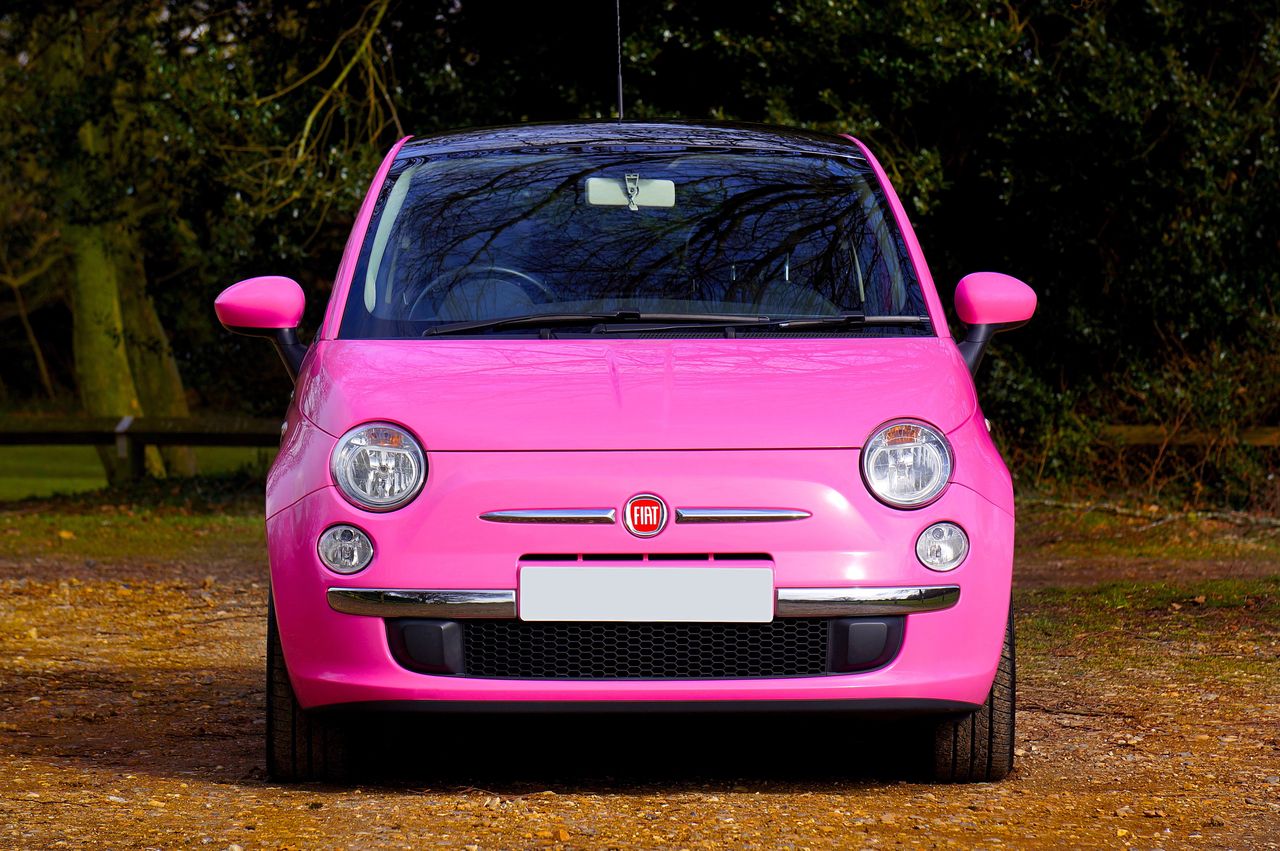 A vibrant pink Fiat 500 car parked outdoors surrounded by greenery and trees.